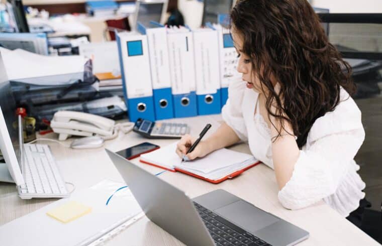 Woman writing in a notebook next to her laptop, working as a freelance writer from home